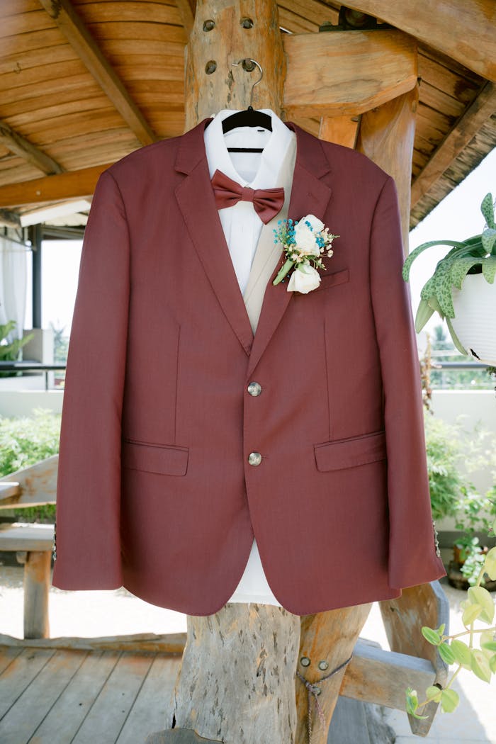 about-us Close-up of a stylish burgundy suit with floral boutonniere hanging on a wooden post.