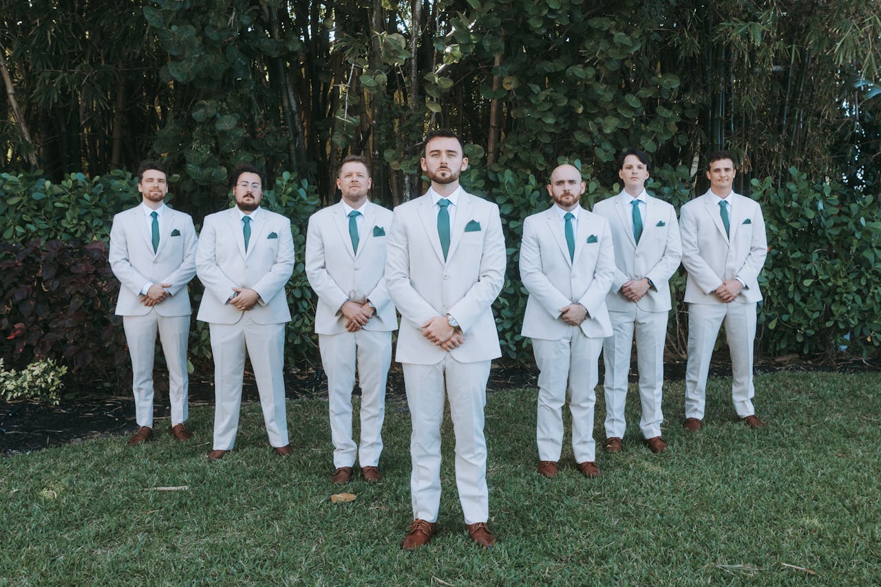 services-05 Seven groomsmen in white suits stand in a garden setting.